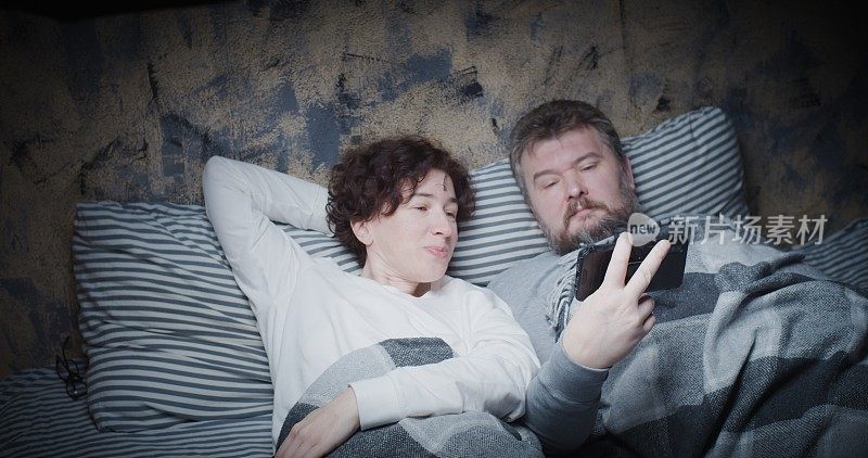 Mature couple is watching video by phone in bed